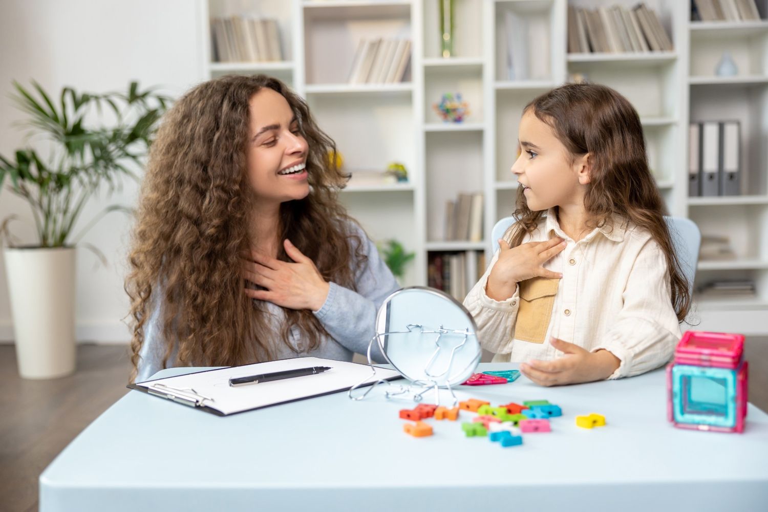 A warm behaviour therapist in Singapore engages with a young girl, both touching their chests, possibly for emotional or speech therapy, with a small mirror and colorful blocks on the table.
