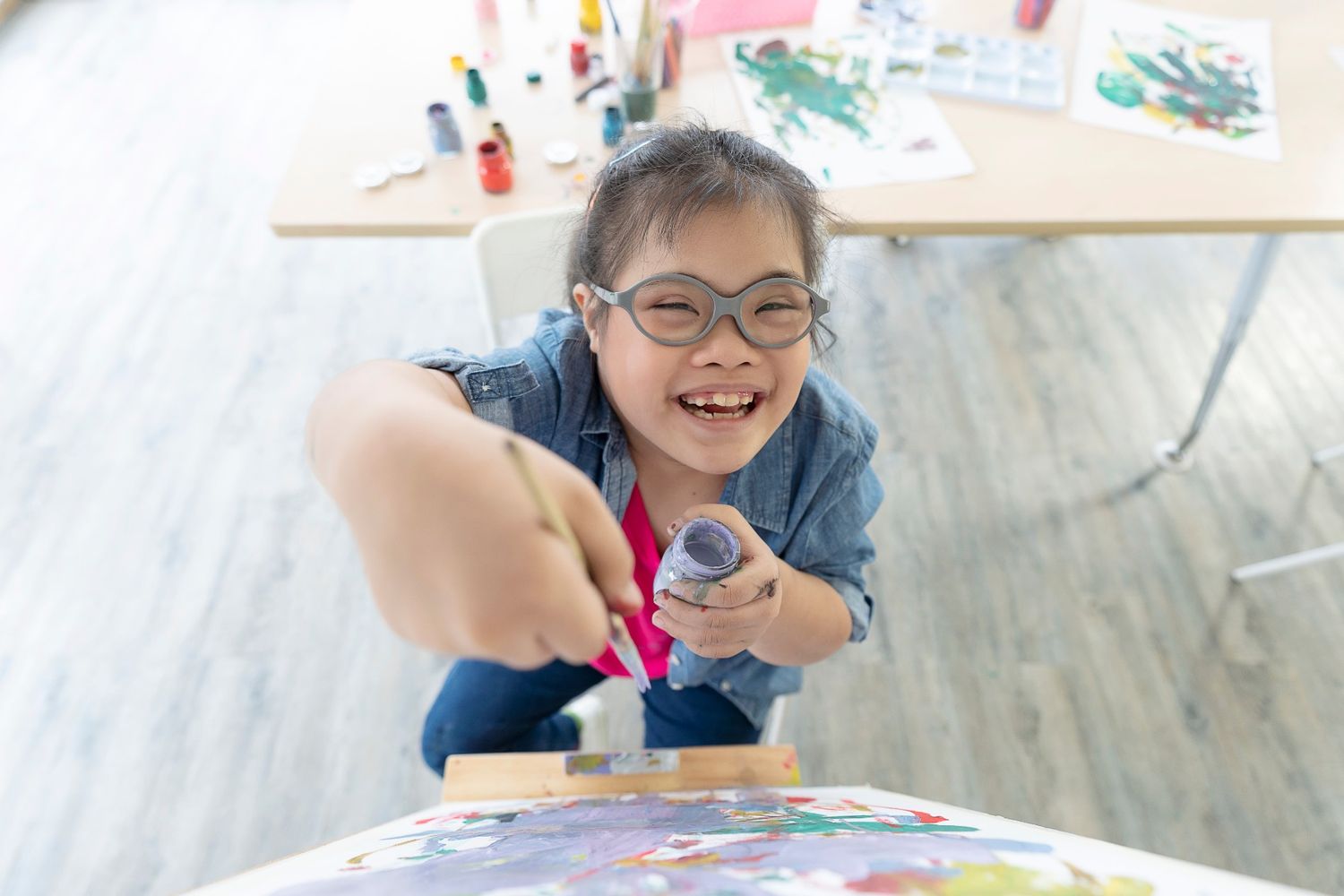 A young girl with glasses joyfully painting on a canvas during an occupational therapy for children session, highlighting play-based learning and fine motor skill development.